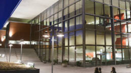 Universitetsbyggnad med stora glaspartier som lyser upp området en mörk kväll.